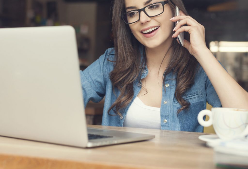 Happy woman talking by mobile phone and using laptop at cafe